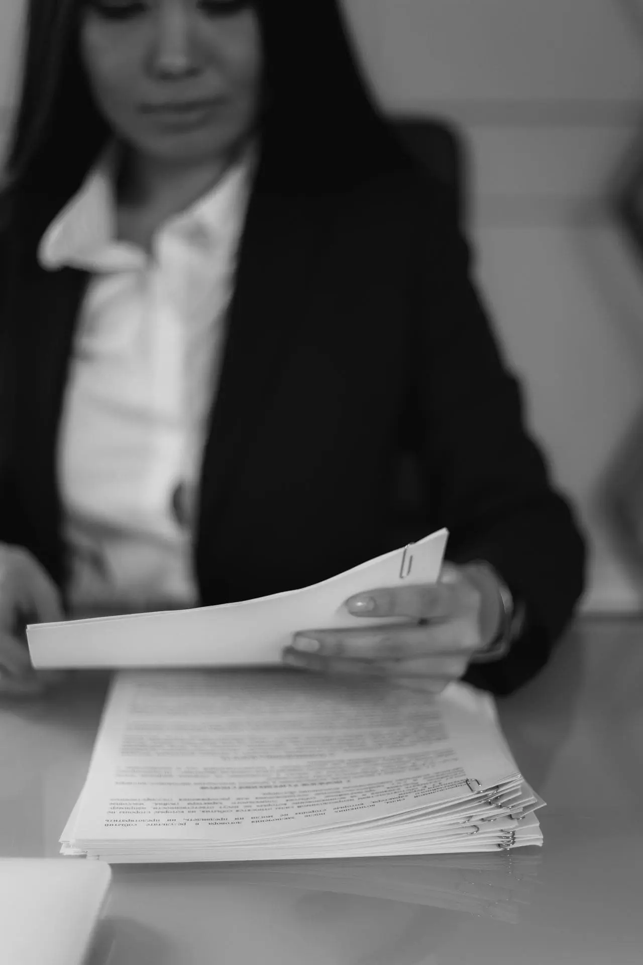 Frau im Anzug liest und hält ein Dokument über einem Stapel Papier auf einem Tisch.