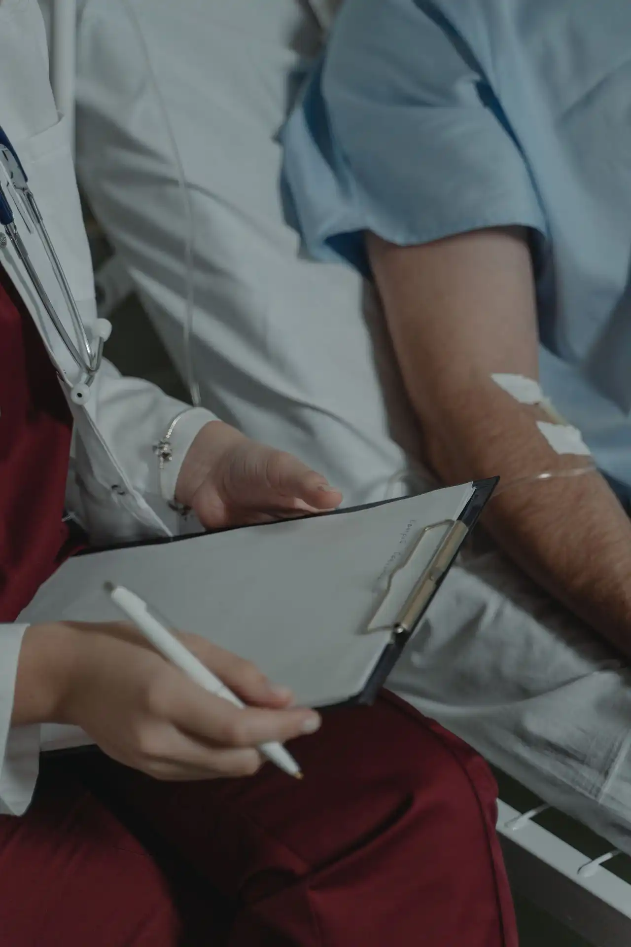 Medizinischer Fachangestellter in Weiß hält Klemmbrett, neben Patienten mit Infusion am Arm im Bett.