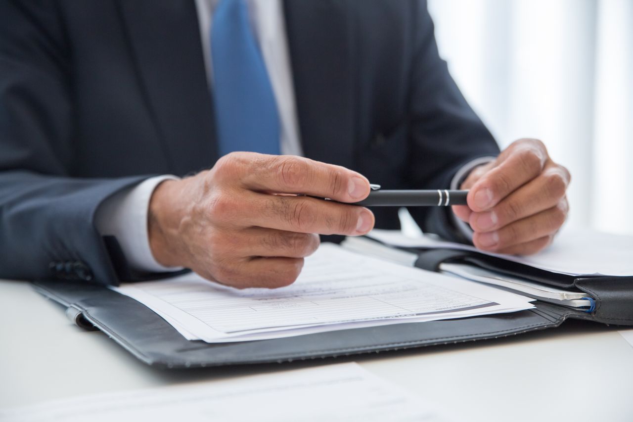 Person in business suit holding a pen over documents on a desk with a black folder