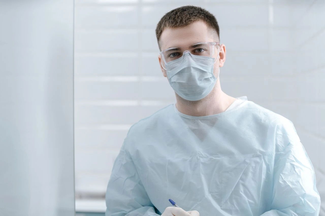 Mann trägt Schutzbrille, Maske und Kittel in einem medizinischen oder Laborumfeld.