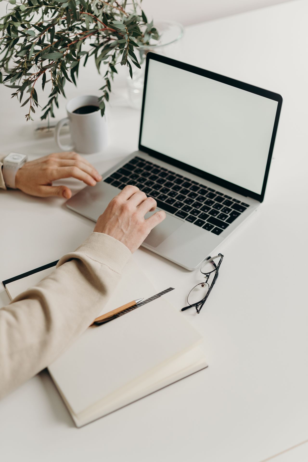 Eine Person arbeitet an einem Laptop mit Leerbildschirm, neben einem Notizbuch, Brille und Kaffeetasse.