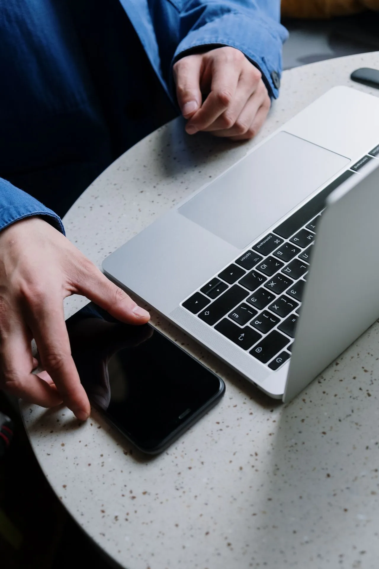 Person in blue shirt reaching to pick up a smartphone next to an open laptop on a speckled table.