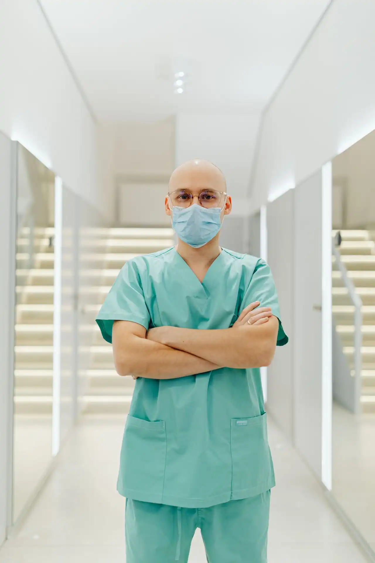 Männlicher Arzt mit OP-Kleidung, Maske und Brille steht mit verschränkten Armen im hellen Flur.