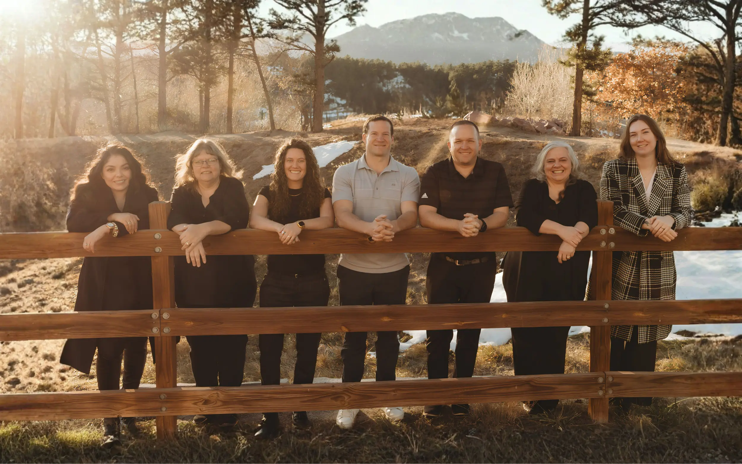 Seven members of the Advanced Dental & Implant Center team smiling and leaning on a wooden fence rail, with a sunlit forest and snow patches in the background