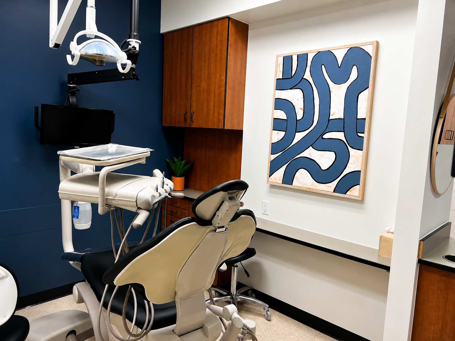 Advanced Dental & Implant center's treatment room featuring a dental chair, overhead exam light, digital monitor, wood cabinetry, and contemporary artwork, creating a clean and welcoming clinical environment.