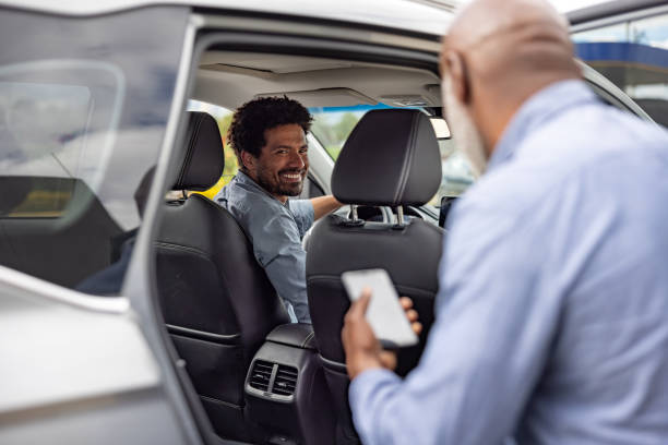Man smiling in driver's seat of car as another man approaches holding a phone.
