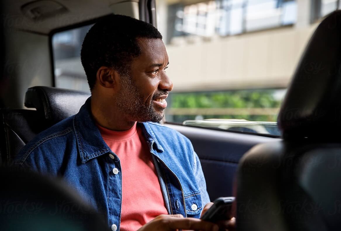 Smiling man in denim jacket holding phone, looking out car window.