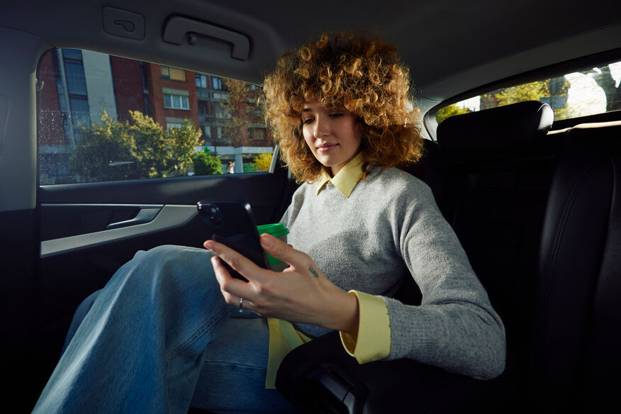 Curly-haired woman in gray sweater sitting in a car backseat, looking at her phone and holding a cup.