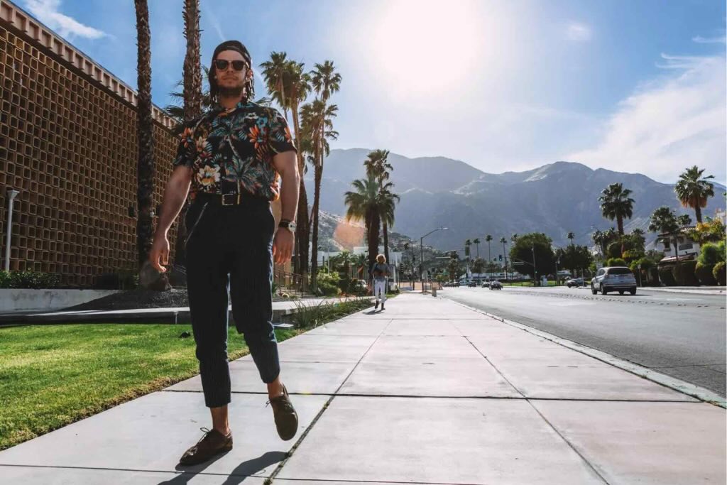 Man wearing sunglasses and a floral shirt walking on a sunny sidewalk lined with palm trees and mountains.