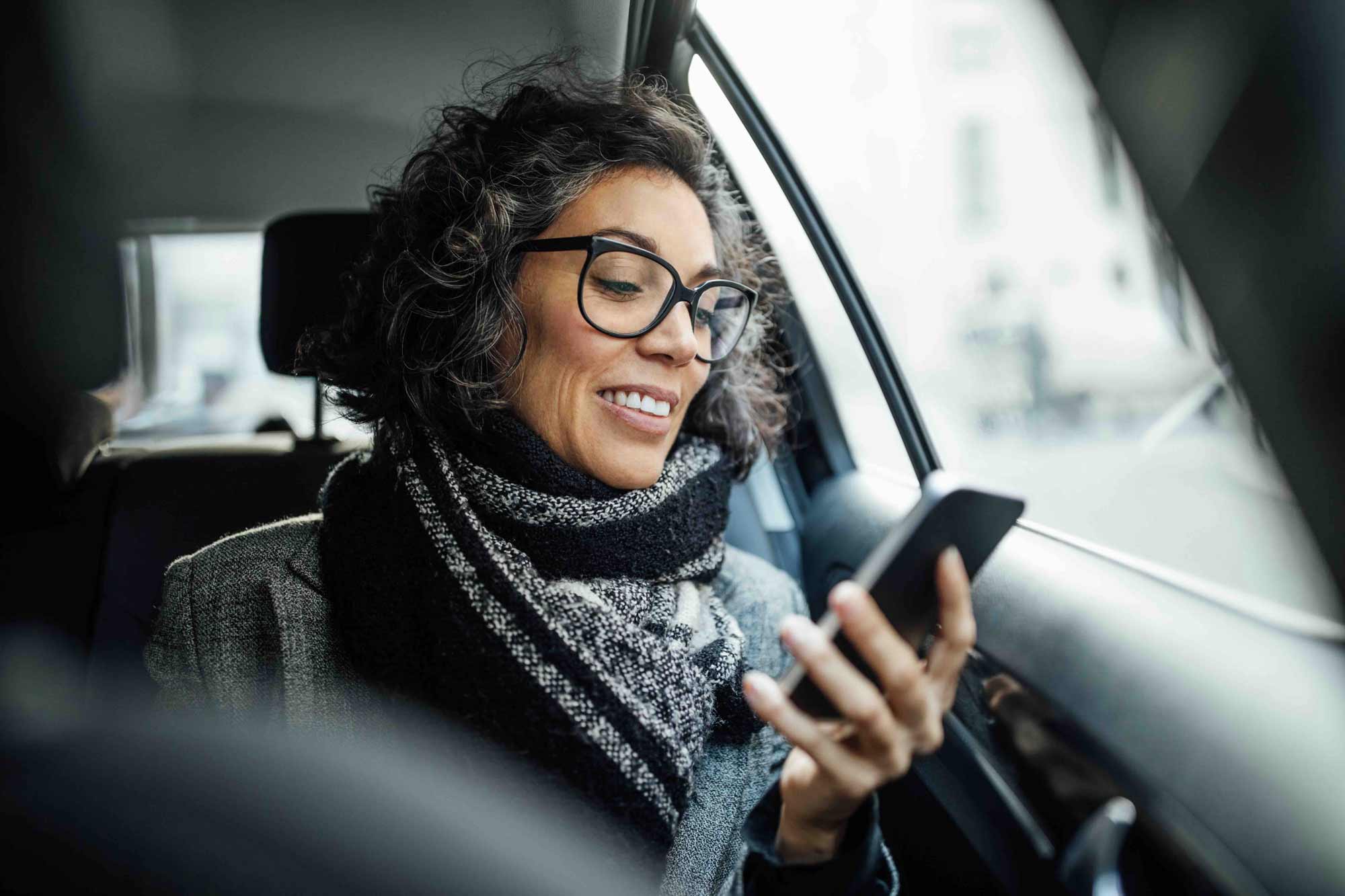 Smiling woman with glasses and scarf looking at her smartphone inside a car.