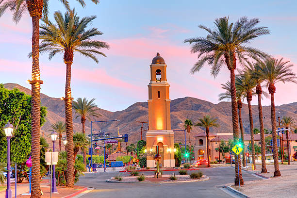 Palm trees and a clock tower lit at sunset with mountains in the background on a town street.