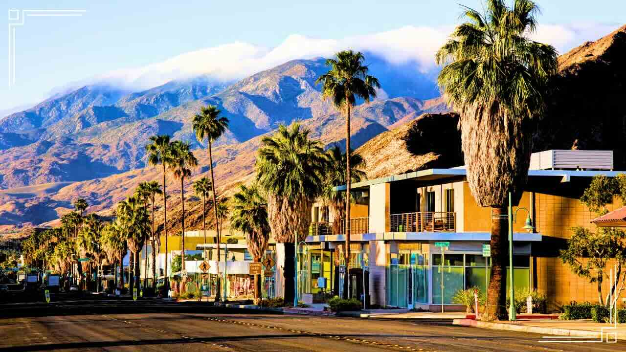 Palm trees line a street with modern buildings under mountains partly covered by clouds.