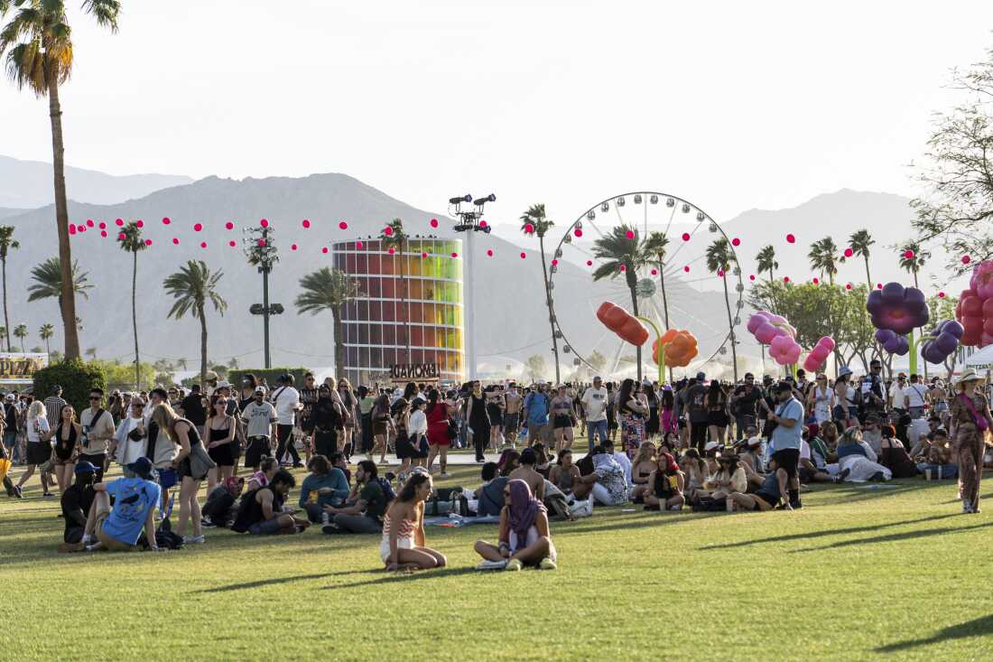 Crowd of people outdoors at a festival with a ferris wheel, colorful balloons, palm trees, and mountains.