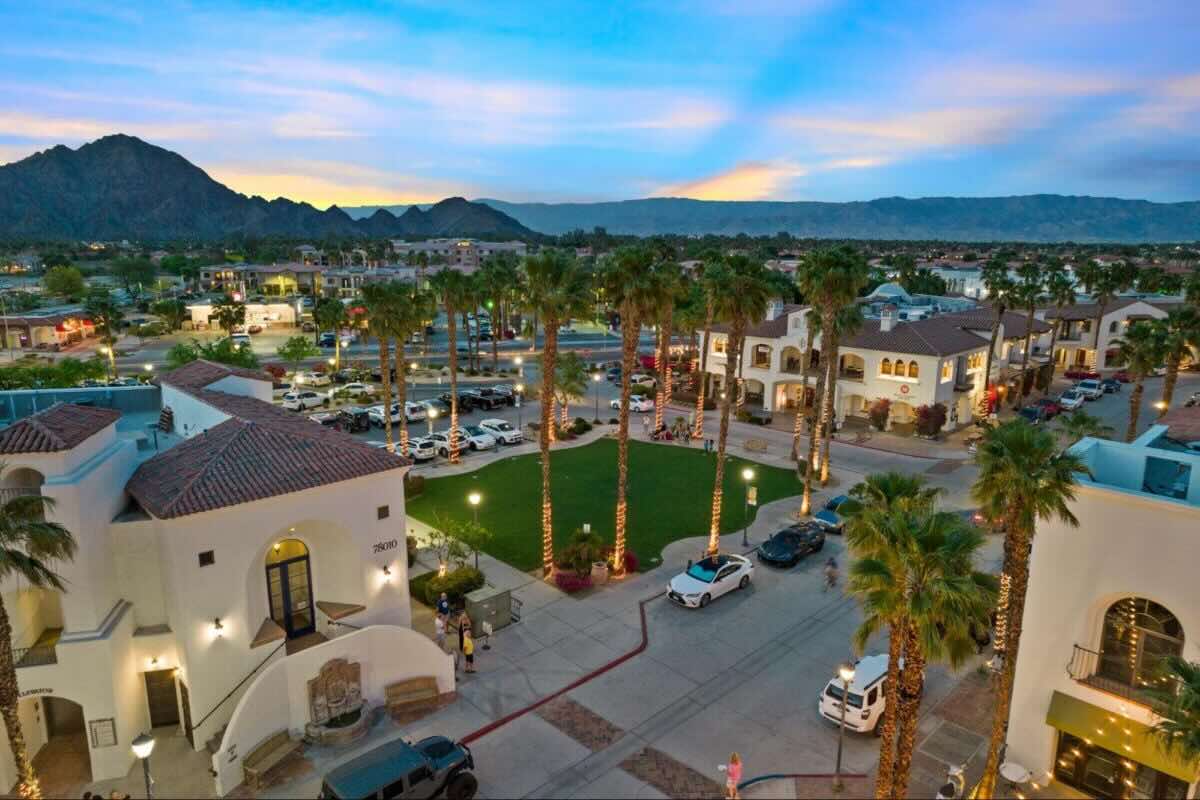 Evening aerial view of a desert town square with palm trees, white buildings, parked cars, and mountains.