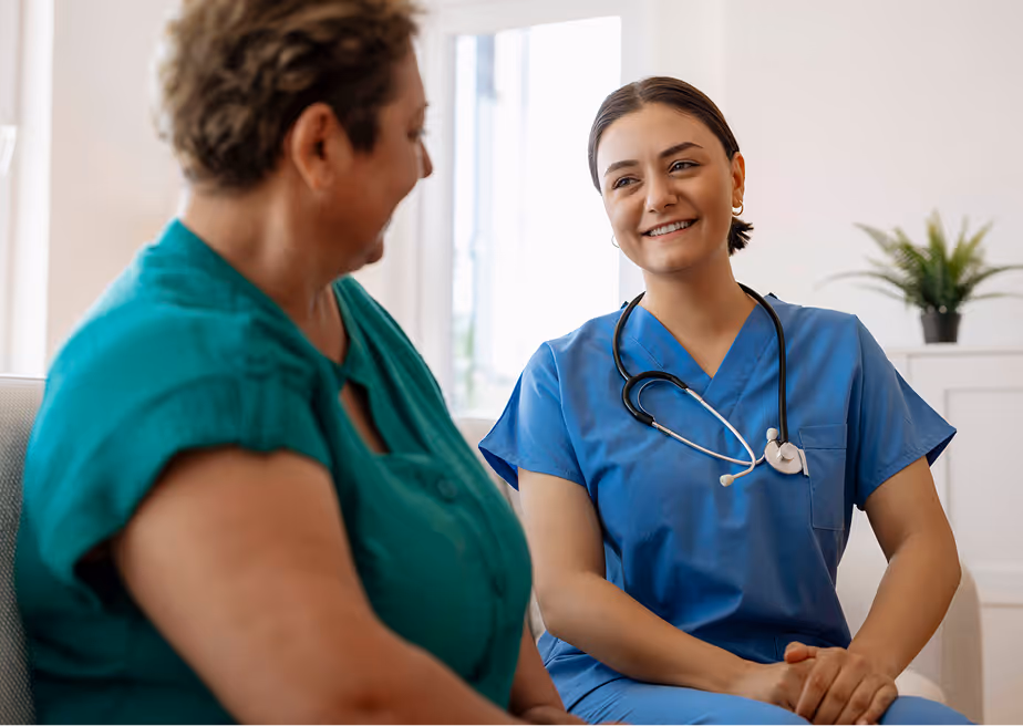 Smiling female nurse in blue scrubs with stethoscope talking to a seated woman in green shirt.