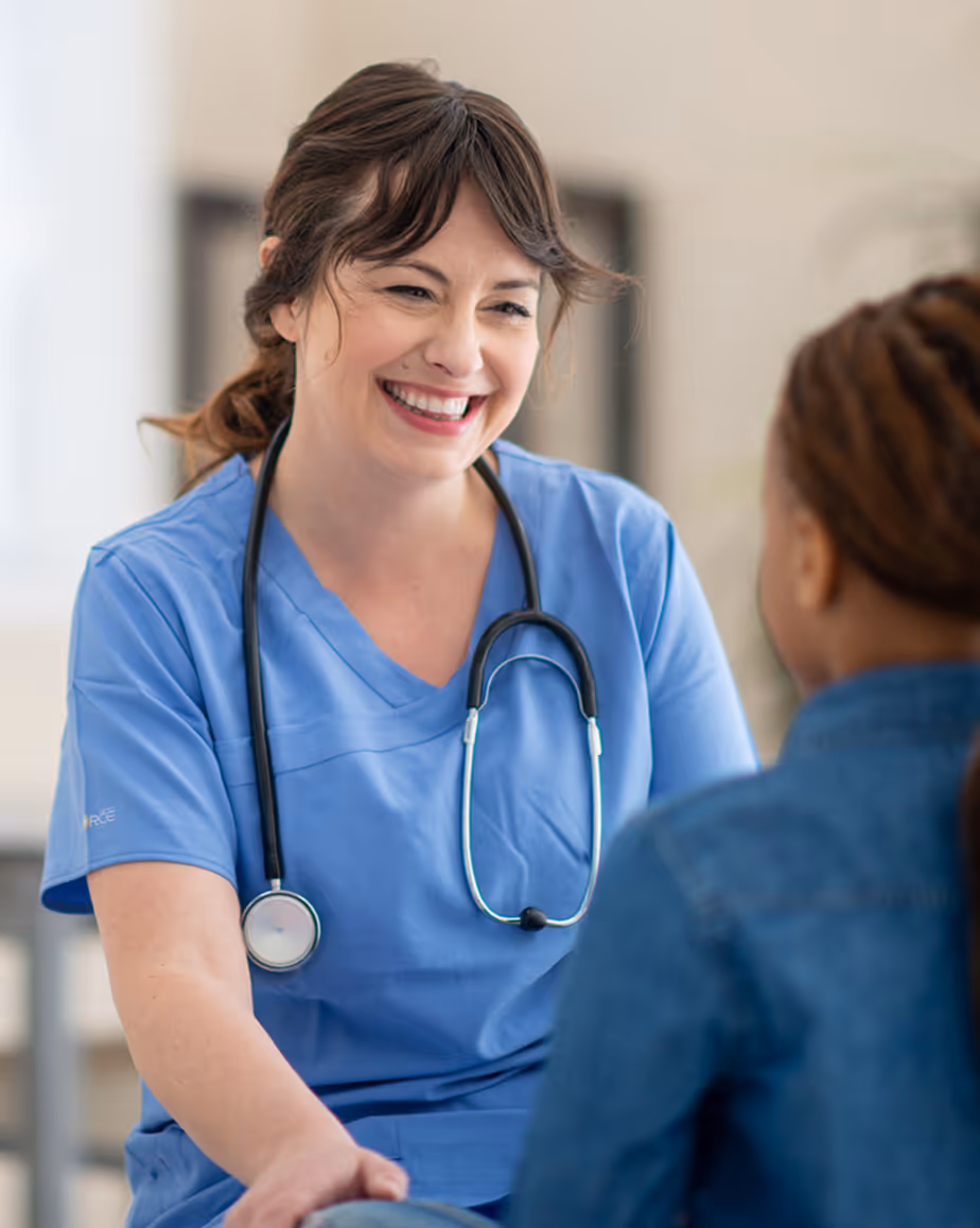 Smiling female nurse in blue scrubs wearing a stethoscope talking to a patient.