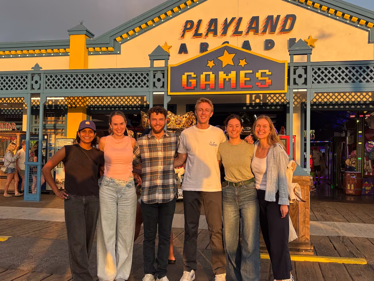 Seven smiling people standing outdoors on a sunny street under beige umbrellas taking a selfie.