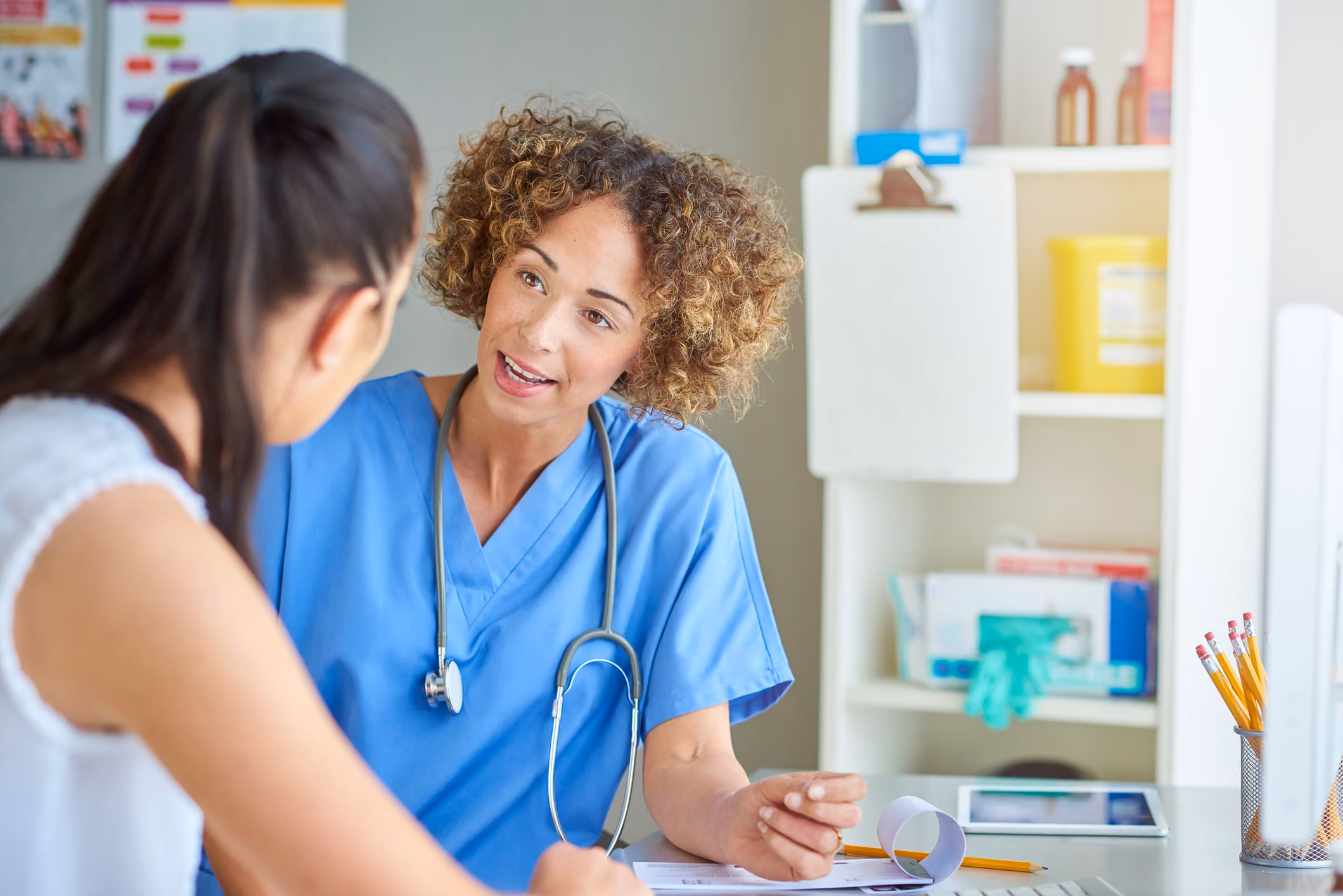 Female doctor in blue scrubs talks attentively with a patient in a medical office.