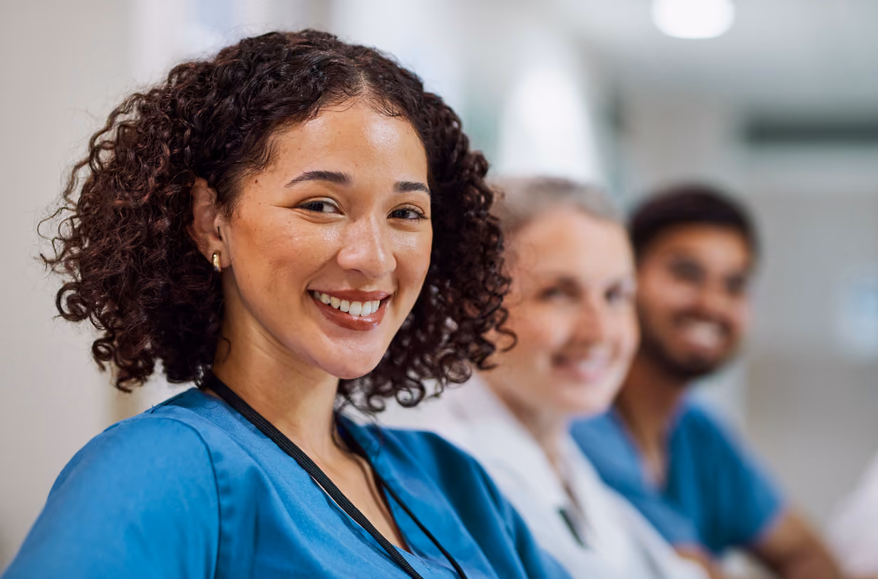 Smiling female healthcare worker in blue scrubs with colleagues in background.