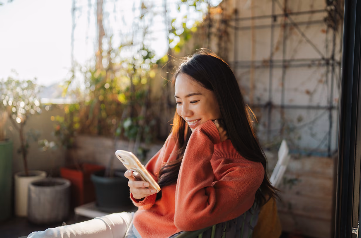 Smiling woman in orange sweater looking at her phone, sitting outdoors near potted plants.