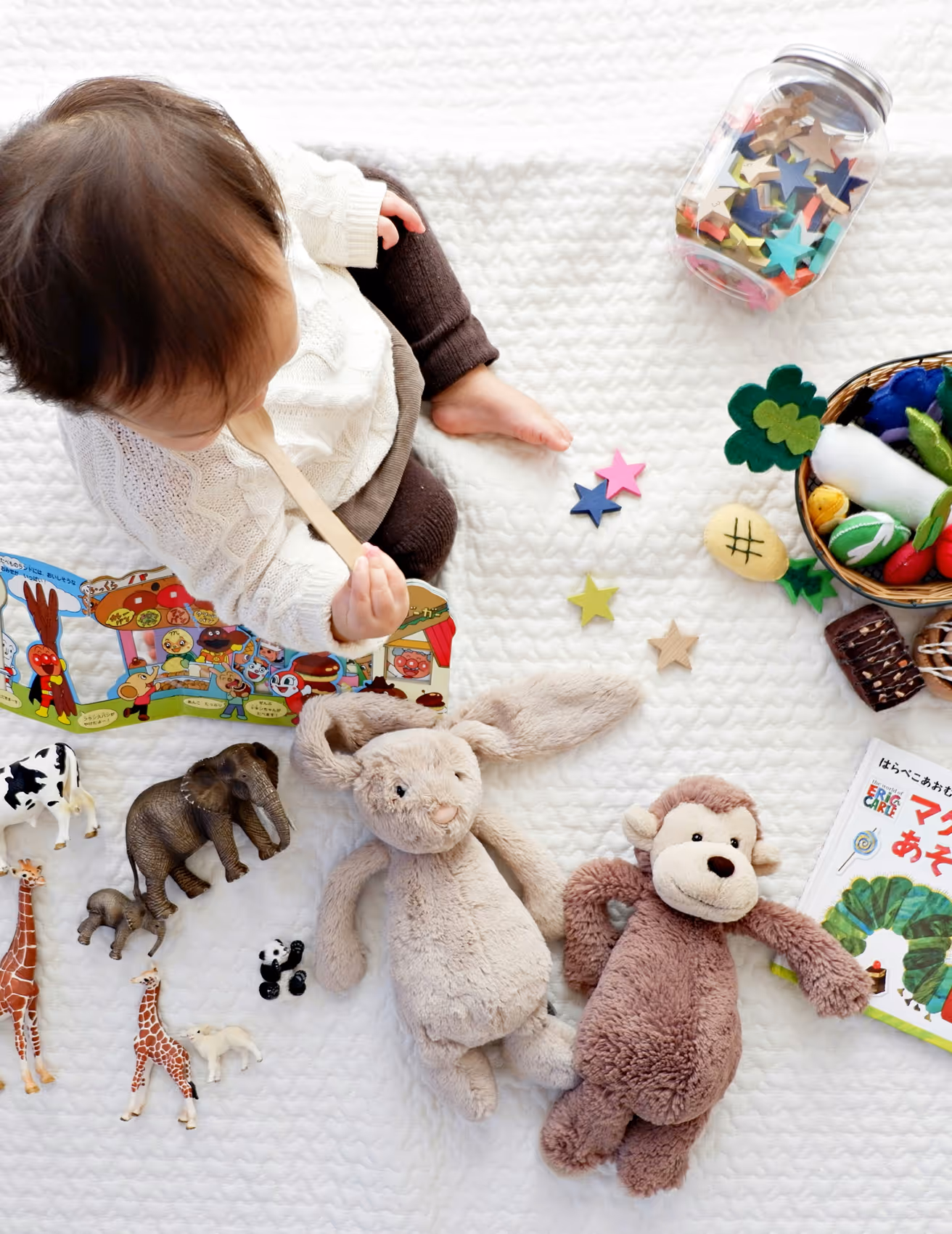 Baby sitting on a white textured blanket surrounded by stuffed animals, toy figurines, colorful wooden stars, and a picture book.
