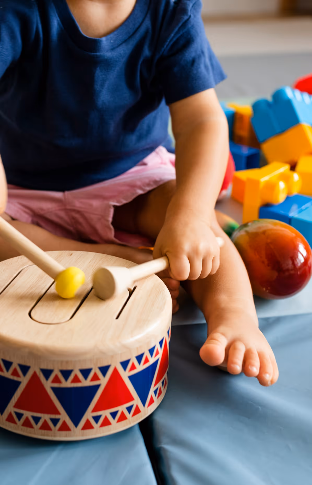 Toddler playing a wooden drum with red and blue triangular patterns while sitting barefoot on a blue mat with colorful toys nearby.