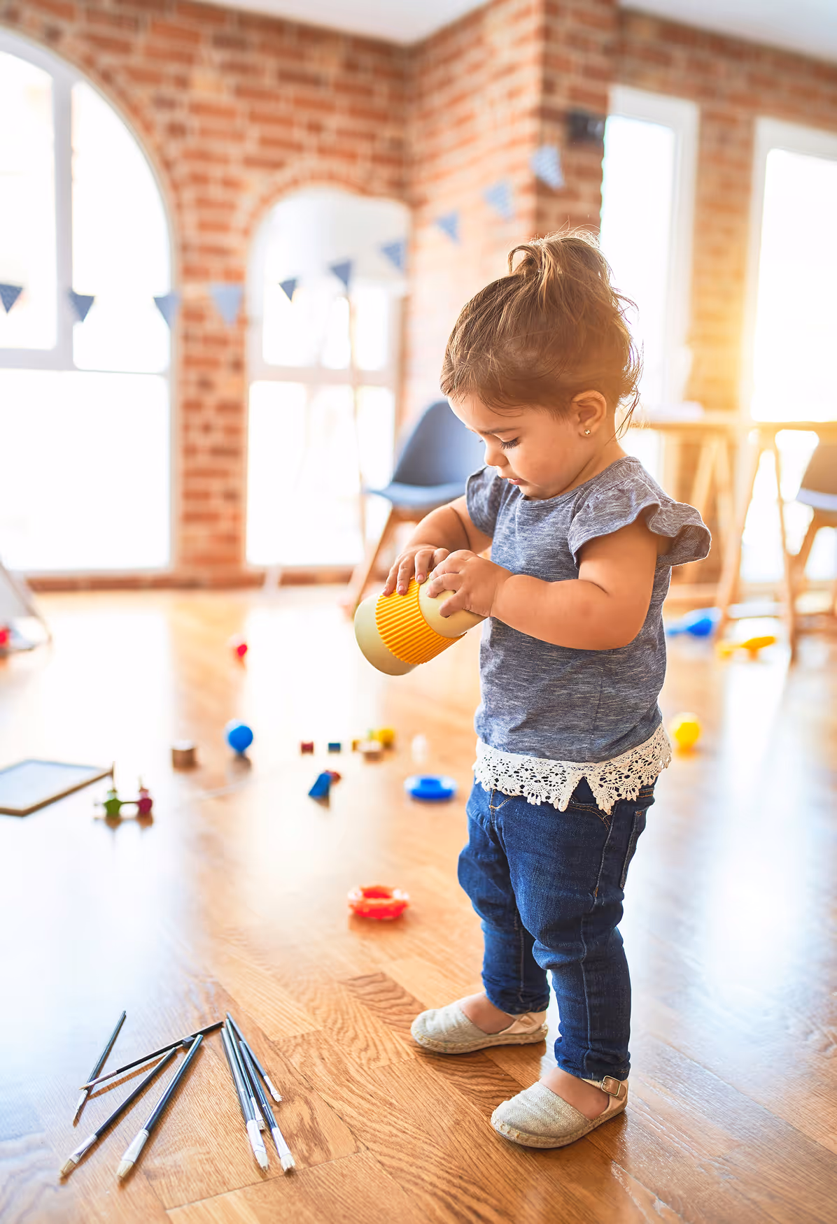 Toddler girl in a blue top and jeans playing with a yellow toy cup on a wooden floor with scattered toys.