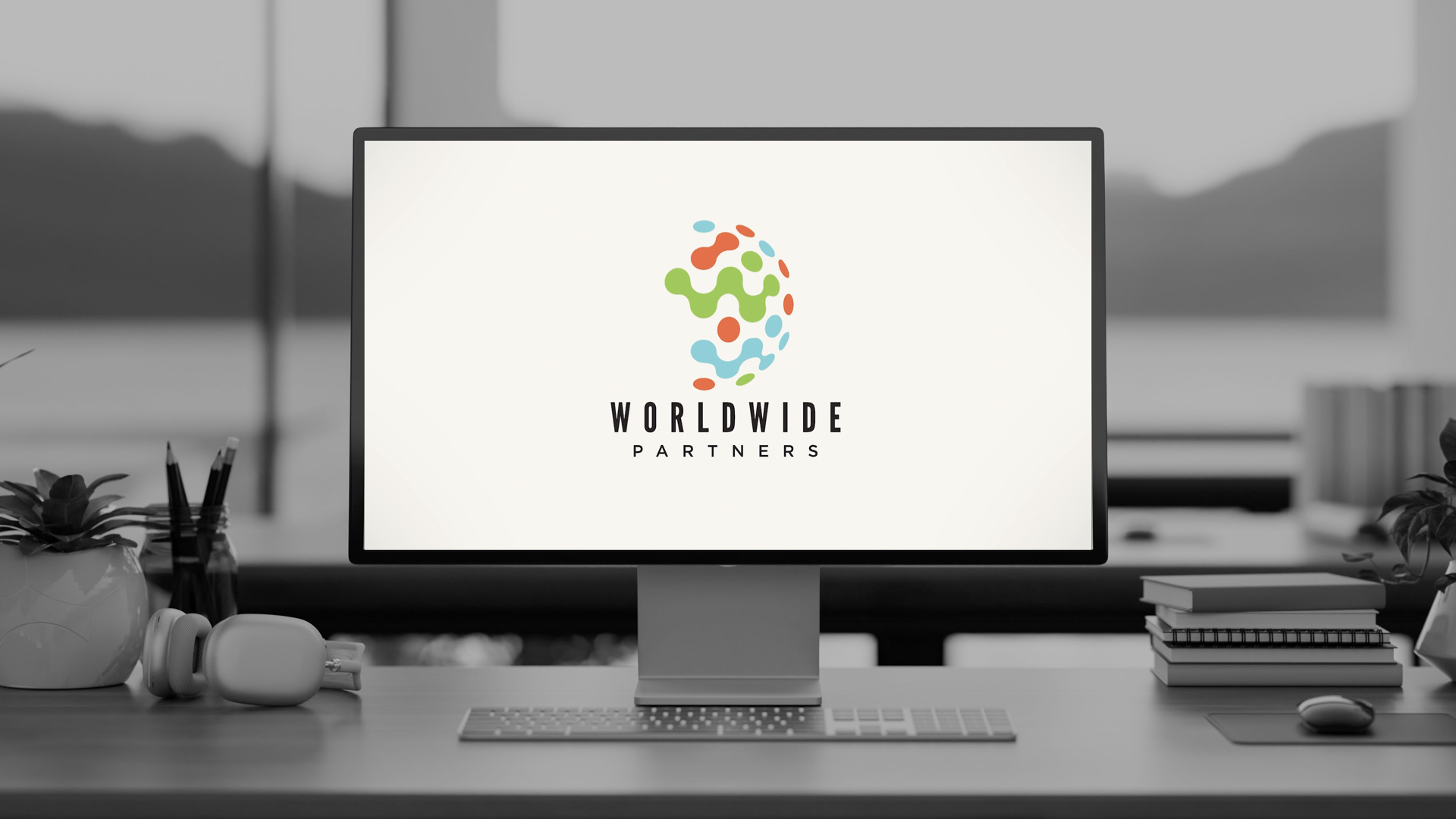 A modern workspace with a computer monitor displaying a colorful abstract globe logo above the text 'WORLDWIDE PARTNERS', a keyboard, wireless mouse, stacked books, potted plants, pencils in a jar, and headphones on the desk.
