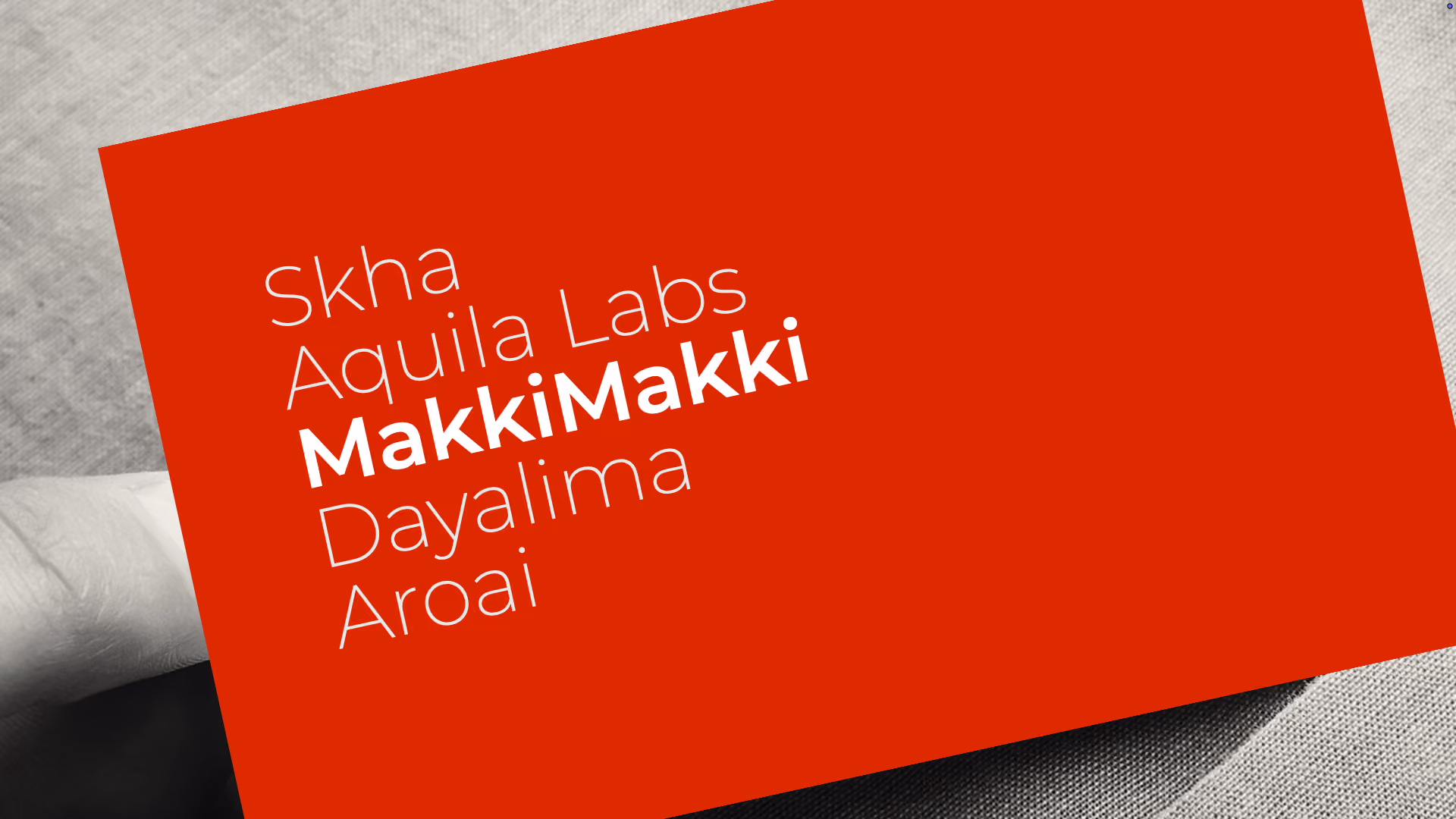 A red card held in a hand with white text listing names: Skha, Aquila Labs, MakkiMakki in bold, Dayalima, and Aroai.