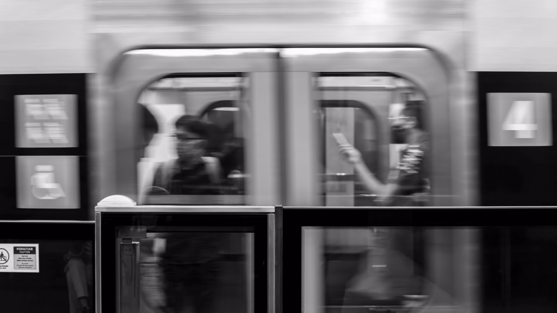 Black and white photo of a subway train in motion with blurred passengers visible through the windows and a platform safety barrier in the foreground.