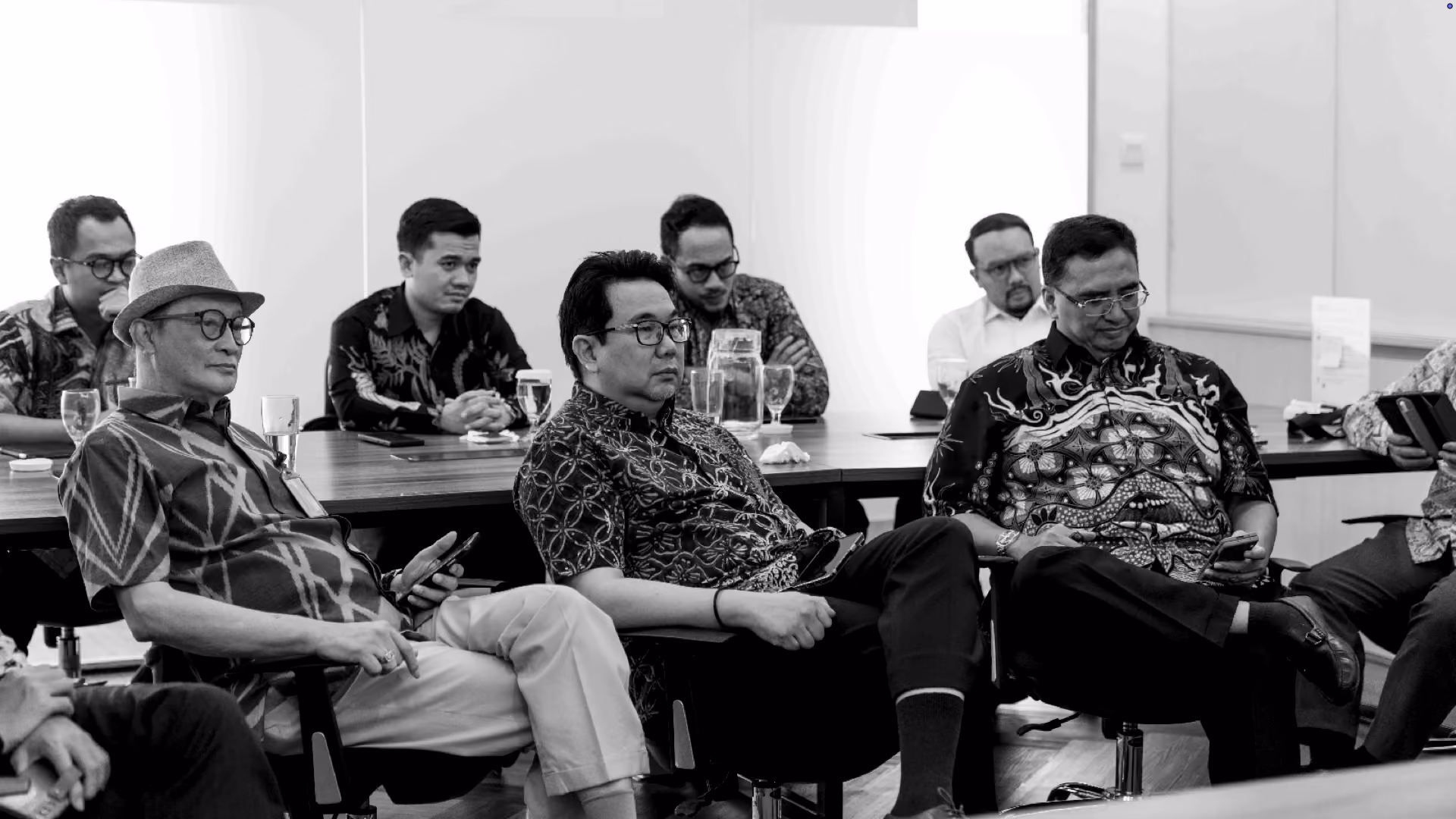 Black and white image of men wearing batik shirts seated in a meeting room, some looking at phones and others attentively listening.