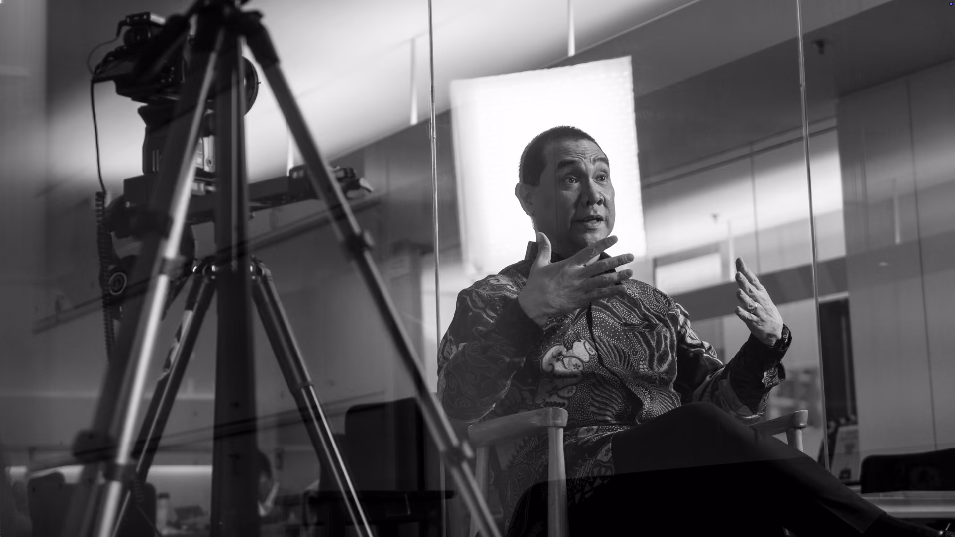 Black and white photo of a man sitting on a chair with arms raised, explaining something during a video recording with a camera setup in front of him in an office setting.