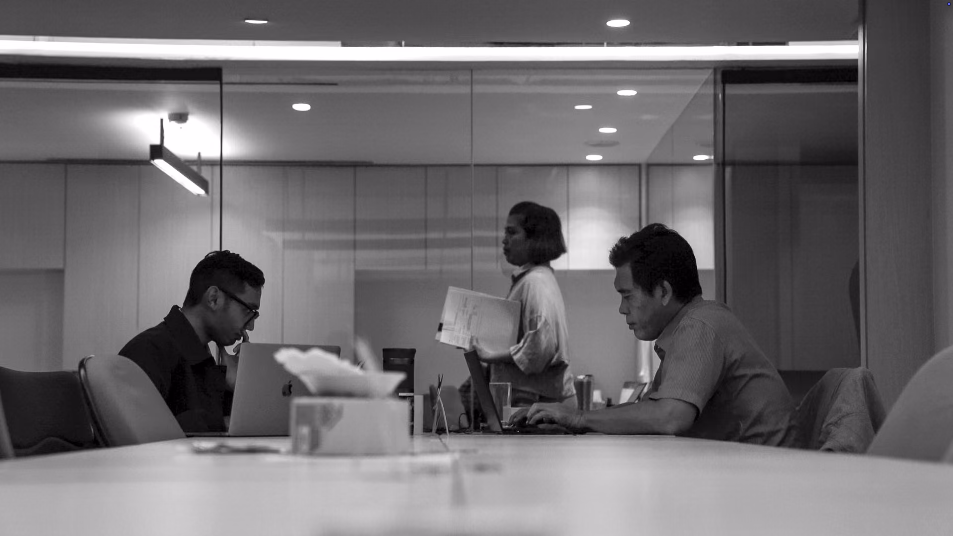 Black-and-white photo of two men sitting opposite each other at a long conference table working on laptops, with a woman standing between them holding papers in a modern office meeting room.