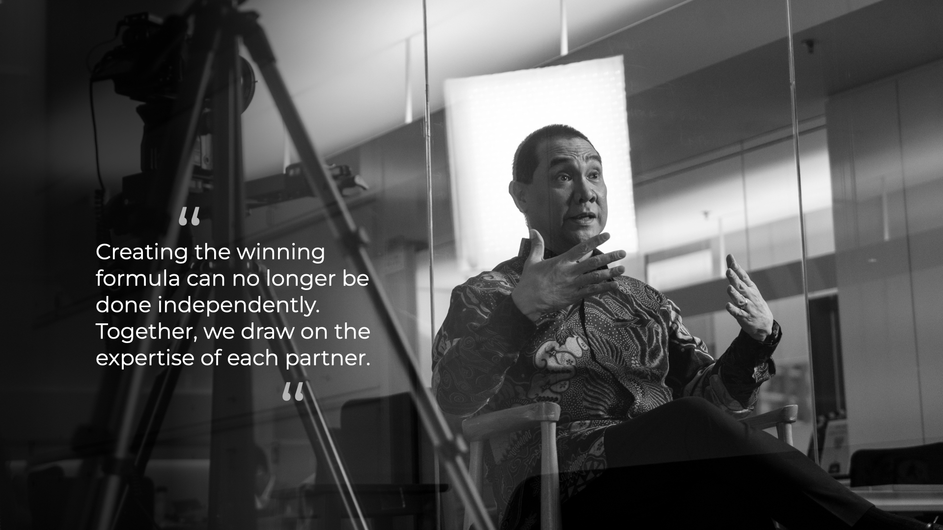 Black-and-white photo of a man in patterned clothing sitting on a chair, gesturing with his hands while speaking in an office setting with a large light panel behind him, and a quote on the left: 'Creating the winning formula can no longer be done independently. Together, we draw on the expertise of each partner.'