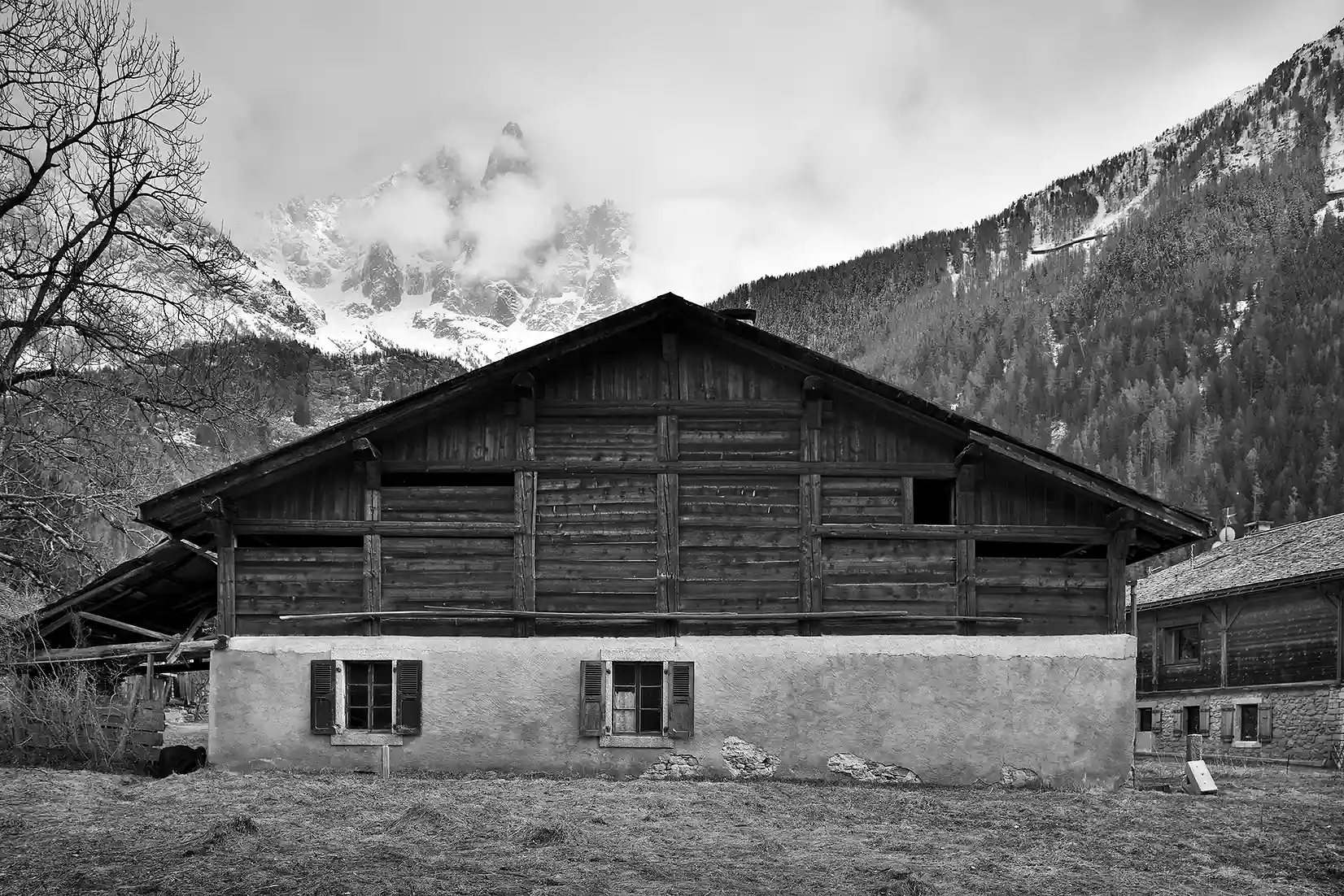 Photo en noir et blanc d'une cabane en bois sur un soubassement en pierre, devant des montagnes boisées et un ciel nuageux, La Ferme des Drus, Chamonix-Mont-Blanc, France.