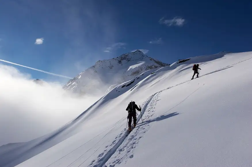 Deux randonneurs équipés de sacs à dos gravissent une pente montagneuse enneigée sous un ciel bleu limpide, La Ferme des Drus, Chamonix-Mont-Blanc, France.