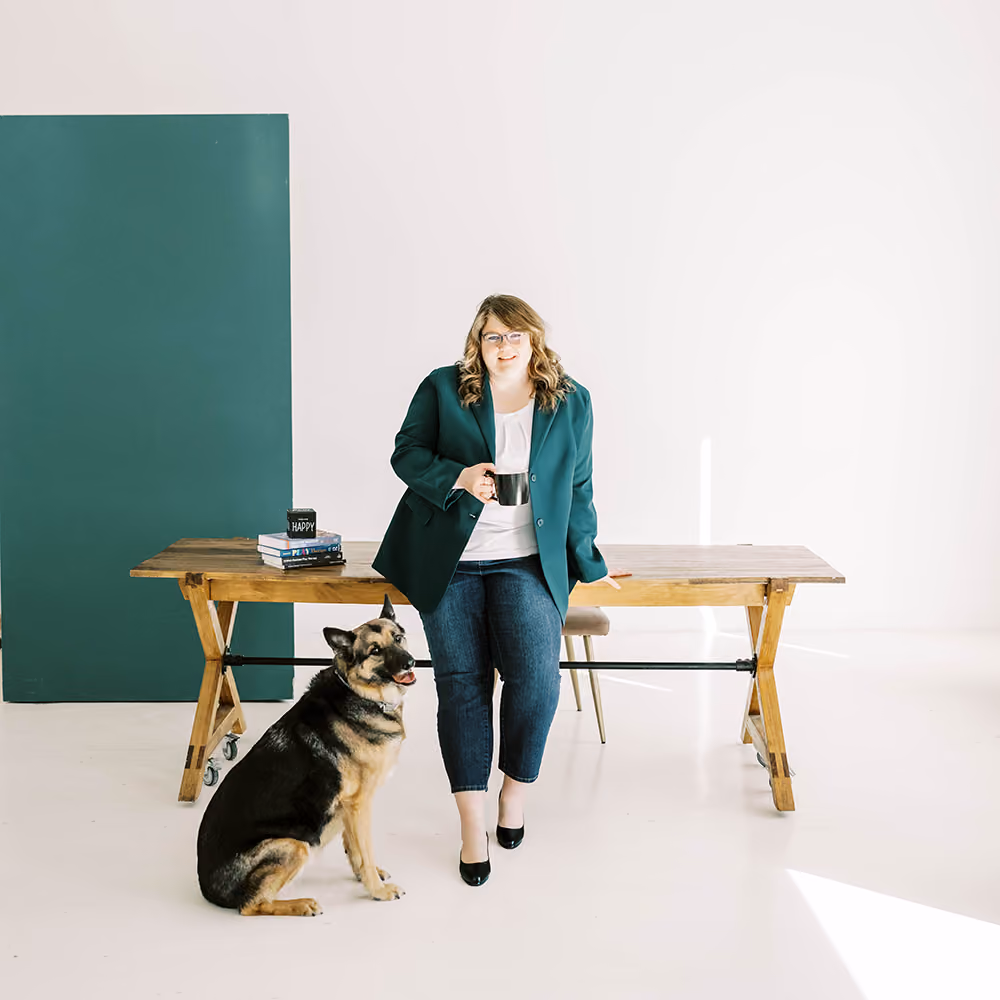 Woman in green blazer sitting on a wooden table with a black mug, beside a sitting German shepherd.
