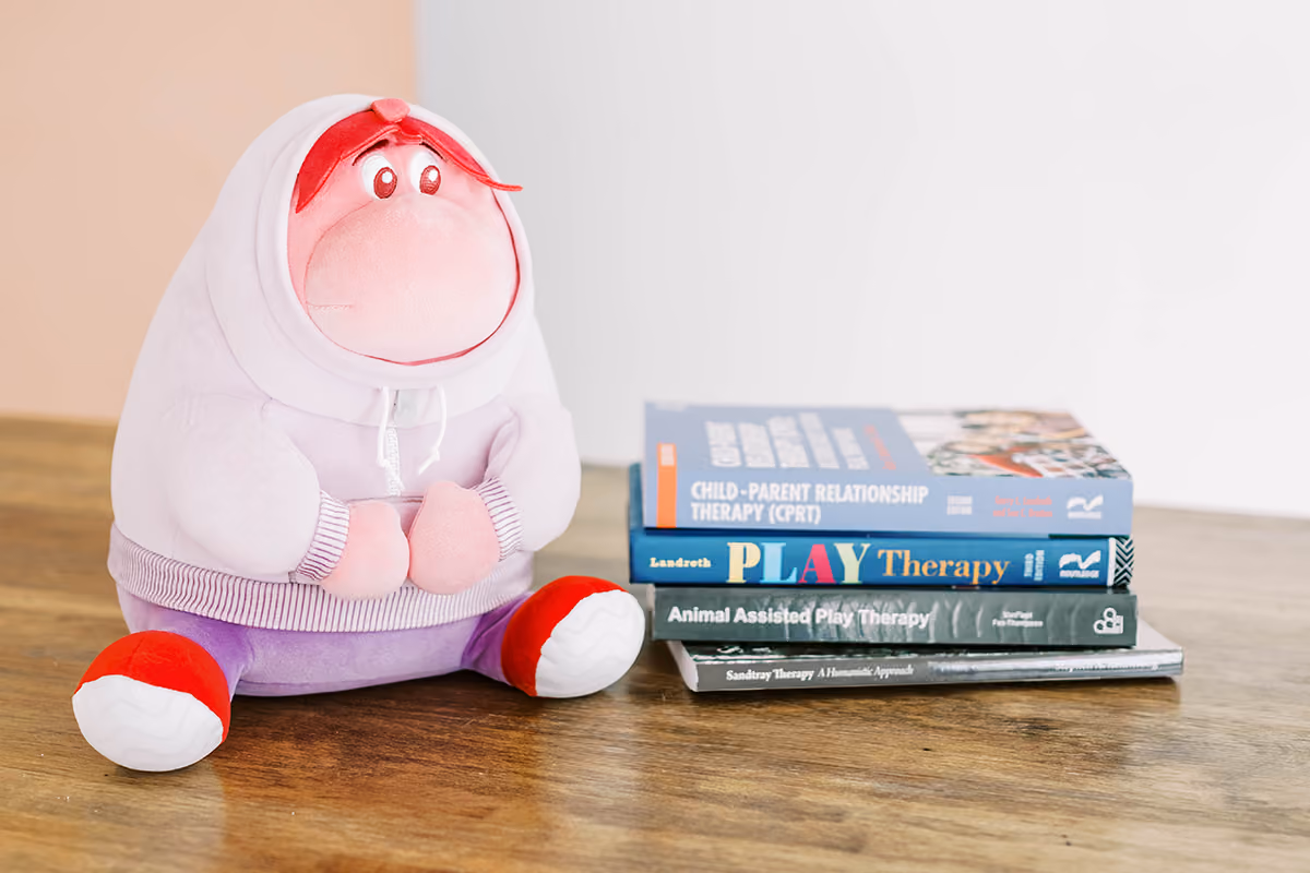Pink plush toy with a hoodie sits next to a stack of therapy books on a wooden table.