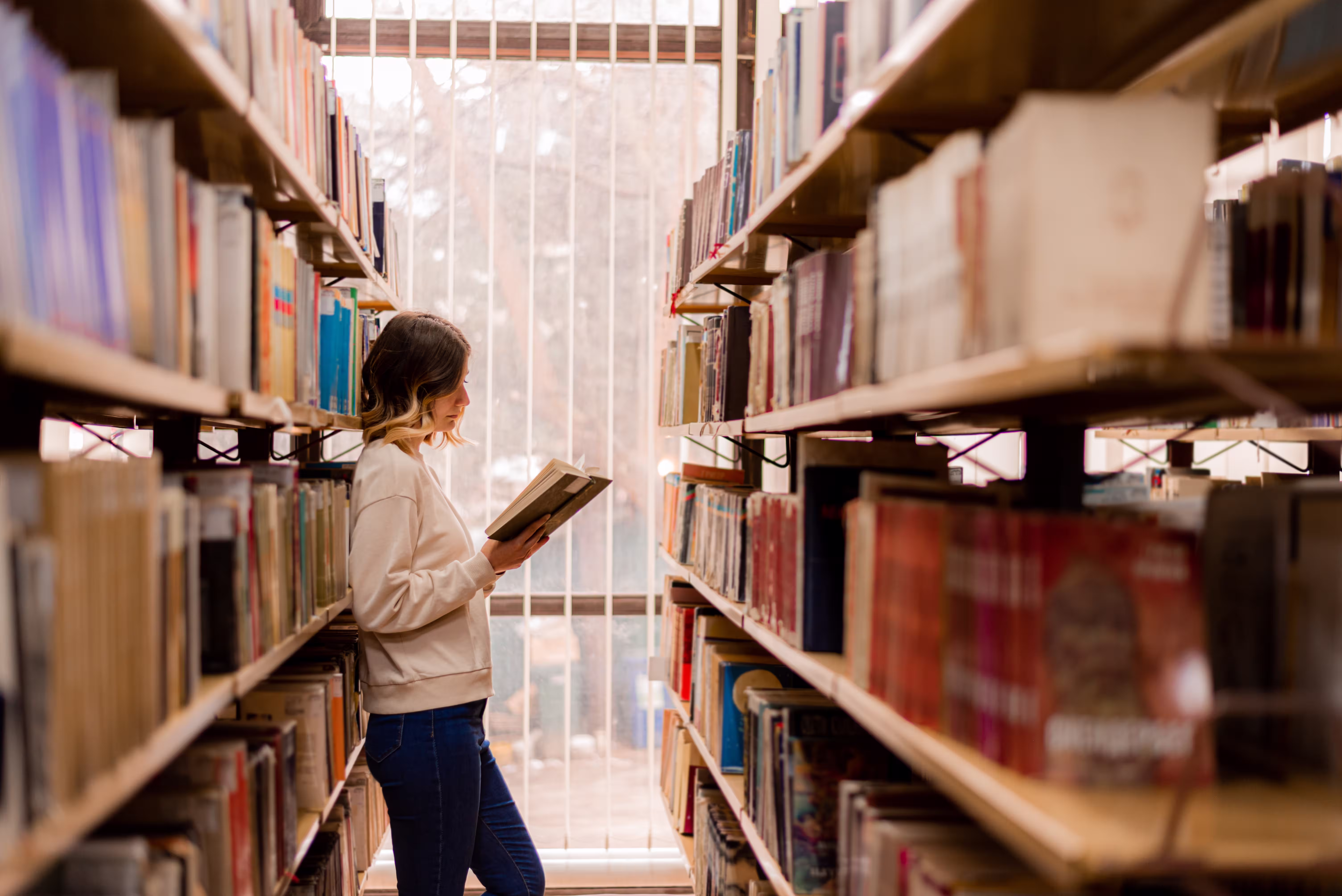 A student standing between library shelves while reading a book and conducting academic research.