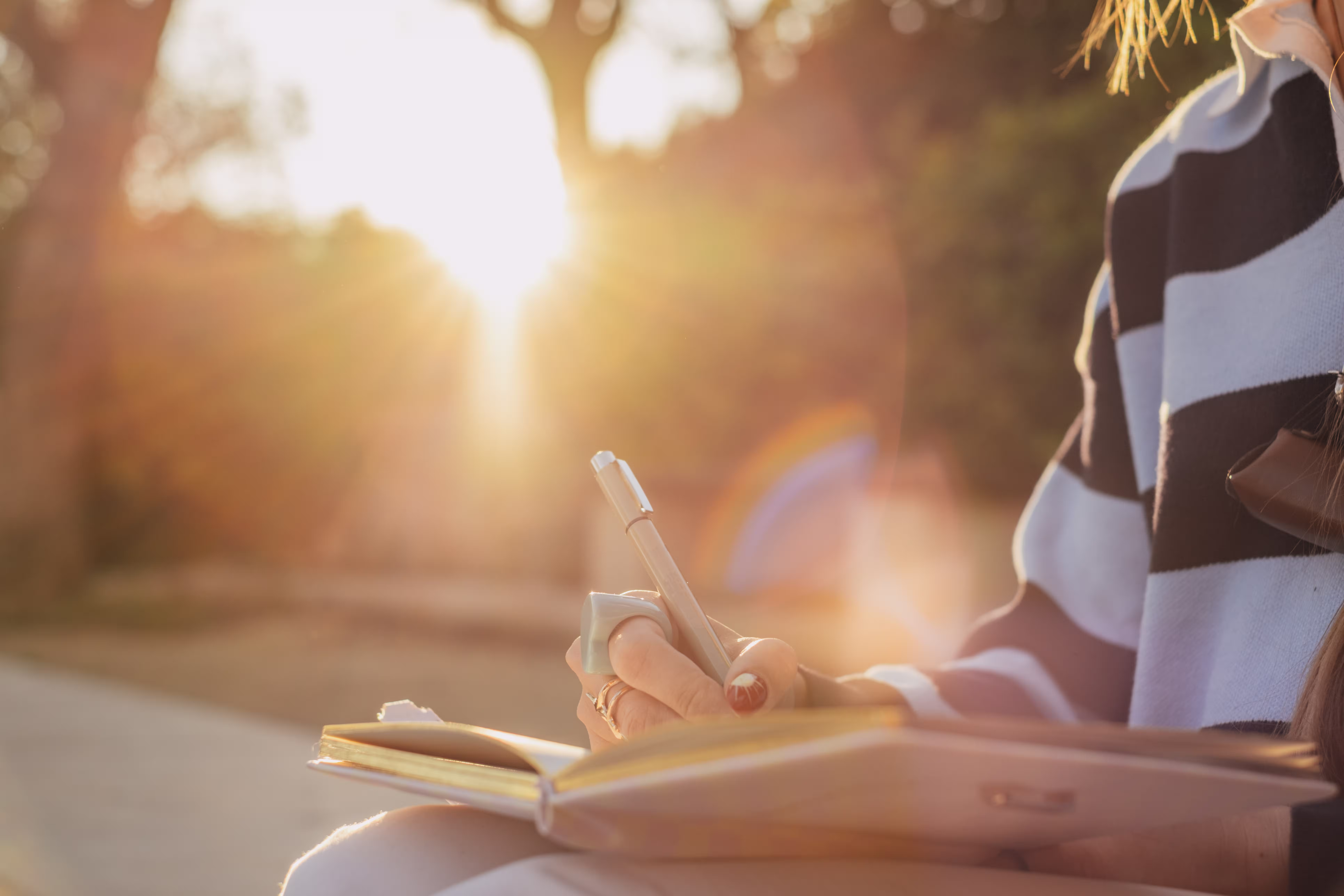 Person writing in a journal outdoors in sunlight, wearing a striped sweater and rings.