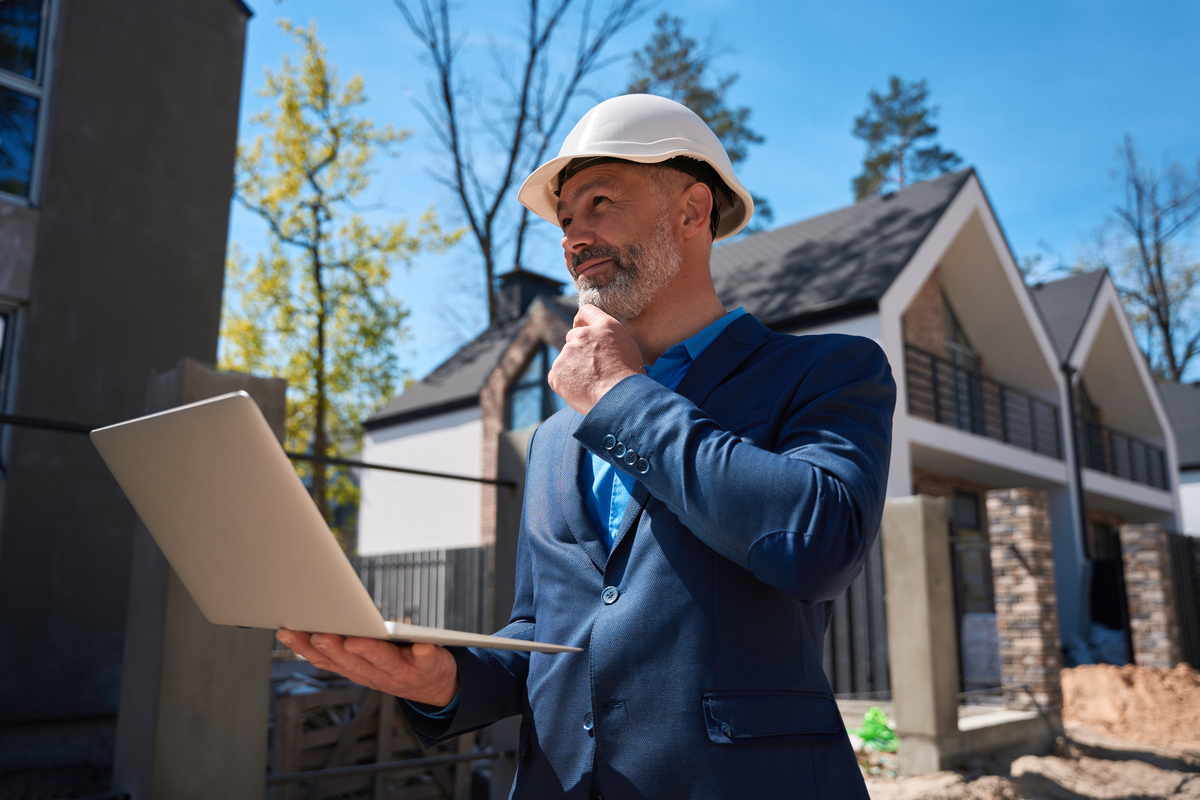 A smiling inspector in a hard hat, thoughtfully looking at a laptop while inspecting a home, symbolizing the wind mitigation inspection process.