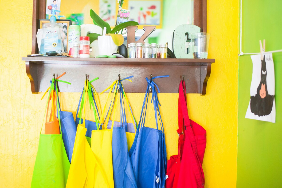 Brightly colored aprons hanging from hooks beneath a wooden shelf filled with various items, set against a vibrant yellow wall in a classroom.