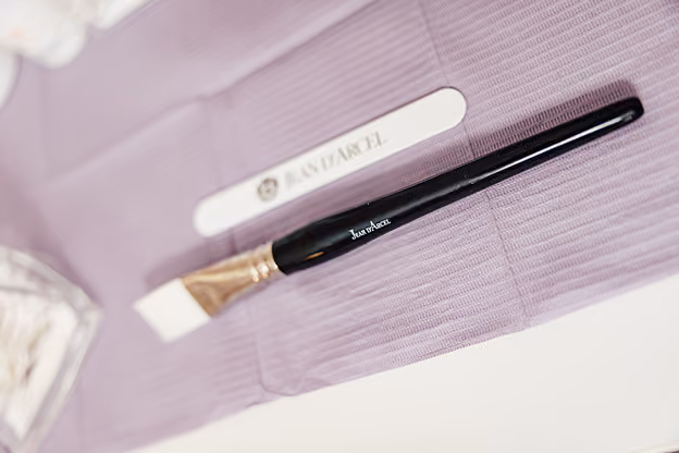 Schwarzer Makeup-Pinsel liegt auf lila Texturuntergrund neben Nagelfeile mit Logo.