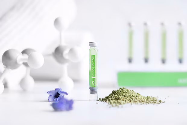 Kleine grüne Flüssigkeit in einem Ampullenfläschchen neben Matcha-Pulver und violetten Blüten.