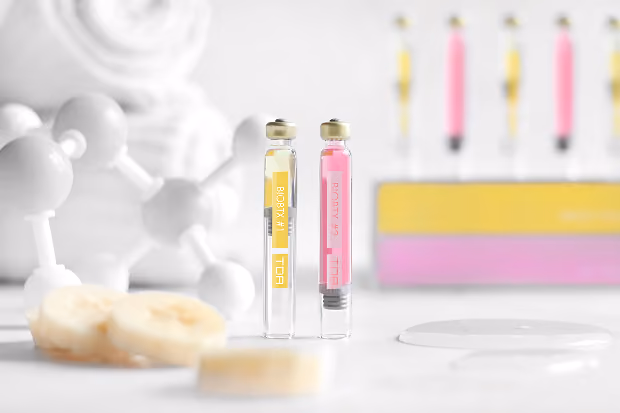 Kosmetikampullen mit gelber und rosa Flüssigkeit, Bananenscheiben und weißen Molekülmodellen.