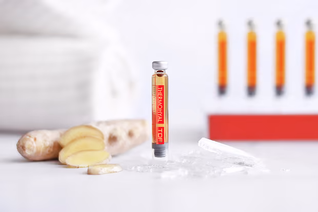 Glasampulle mit rotem Etikett neben Ingwerscheiben und Eiswürfeln auf weißem Untergrund.