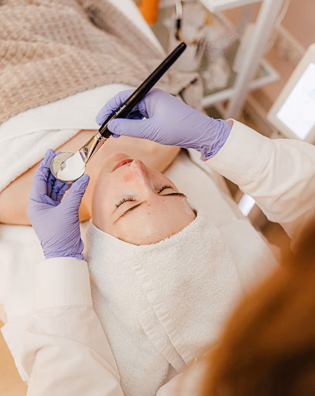 Kosmetikerin in lila Handschuhen trägt mit Pinsel eine Gesichtsmaske auf die Stirn einer entspannten Frau auf.