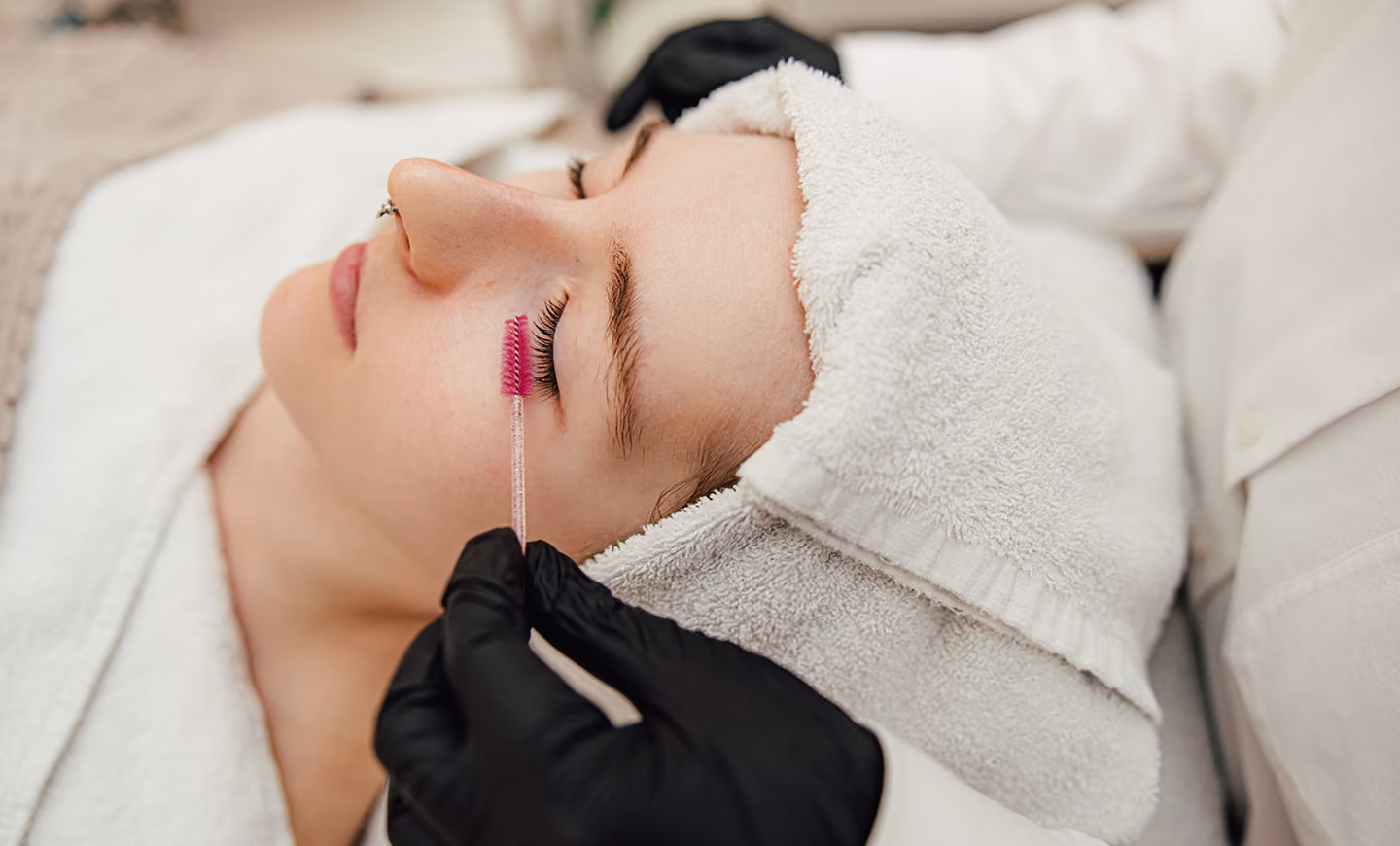 Kosmetikerin trägt Wimpernlifting auf eine liegende Frau mit weißem Handtuch um den Kopf auf.