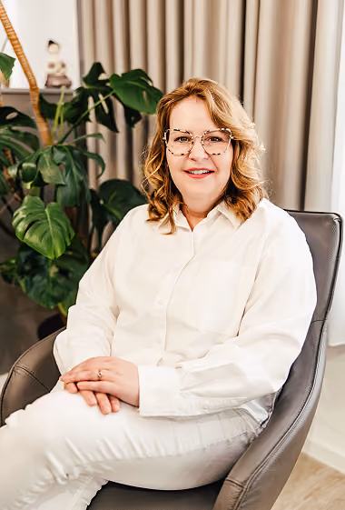 Maike Kahllund sitzt auf einem Stuhl, trägt weiße Bluse, grünes Blatt im Hintergrund.