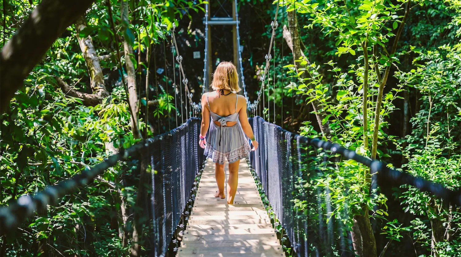 Woman in a blue dress walking barefoot on a rope suspension bridge in a lush forest.